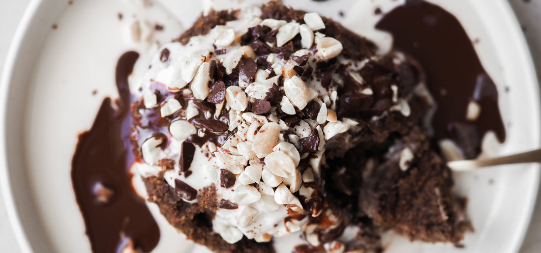 Chocolate Mug Cake s ovsenými vločkami, banánom a vegánskym proteínom, ozdobený rastlinným jogurtom, horkou čokoládou a arašidmi.